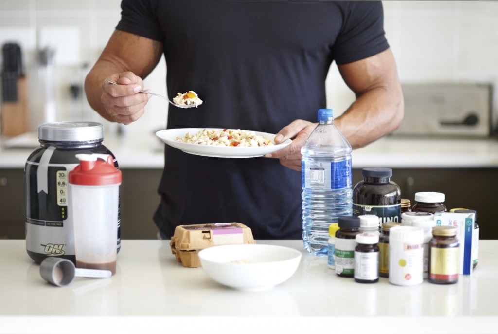 A muscular man holding a bowl of muesli standing in front of his supplements