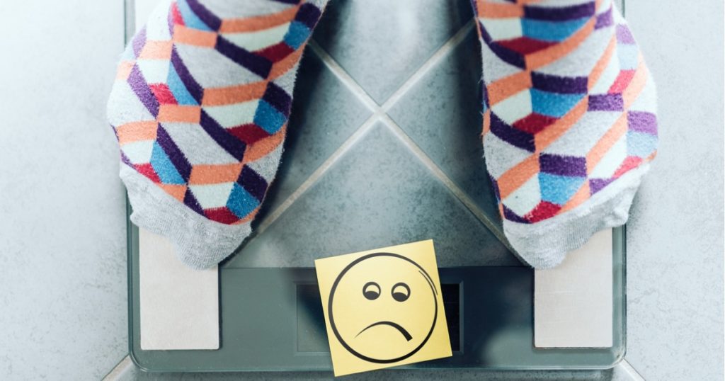 Overhead view of a person in multi-colored socks standing on scale with a yellow sticky note with a sad face covering the number