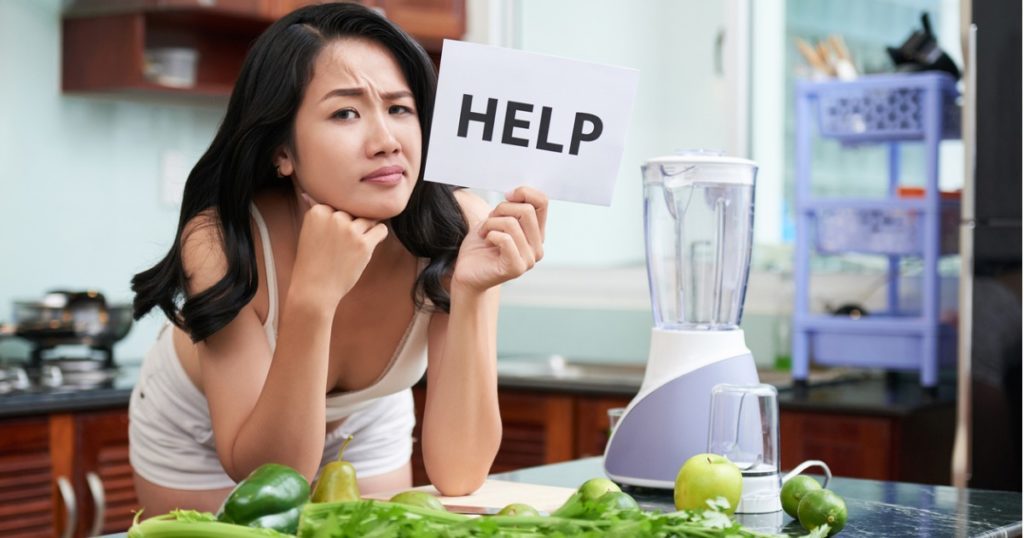 Woman leaning against kitchen counter with fruits and vegetables spread across, holding a white sign that says help.