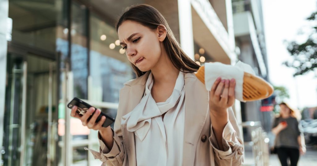 Woman standing outside a building holding a sandwich in one hand and scrolling through her cellphone in the other hand.