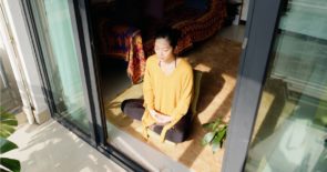 Young asian female sitting cross-legged on the floor in front of an open sliding glass door.