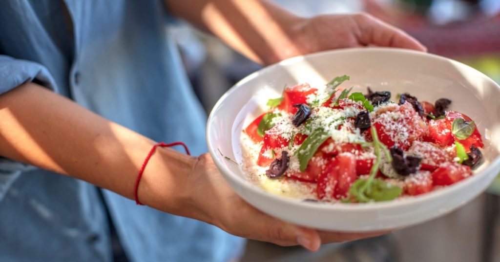 Closeup of hands holding a bowl of sliced tomatoes, olives, and red onions.